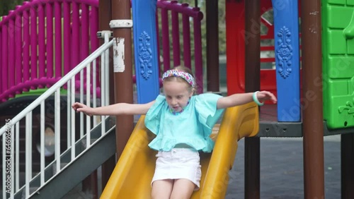 Playful little girl in casual blue clothes rides slide in maze on children playground smiling schoolgirl plays attraction in public park child enjoys weekend