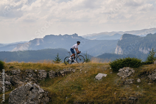 Professional male cyclist is riding a gravel bike on a gravel road with an amazing view of the mountains. Cyclist is practicing on gravel road. Man is riding on top of a hill.Bucegi Mountains, Romania photo