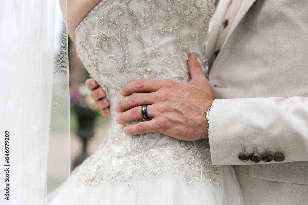 The groom hugs the bride by the waist during the wedding ceremony. Hugs of the bride and groom close-up. Wedding ring on the groom's finger. Beautiful wedding dresses for the bride and groom The groom hugs the bride by the waist during the wedding ceremony. Hugs of the bride and groom close-up. Wedding ring on the groom's finger. Beautiful wedding dresses for the bride and groom
