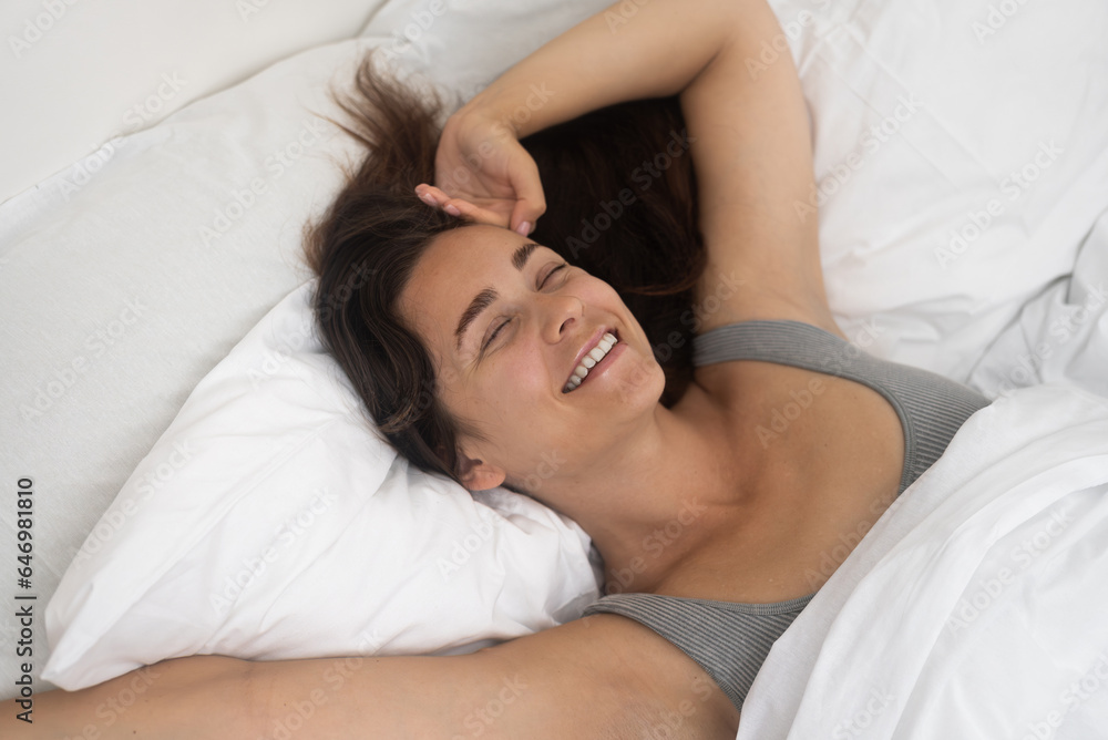 Smiling woman holds hand on forehead resting on bed with soft blanket in accommodation bedroom happy lady energized from sleep enjoys morning lying on mattress Smiling woman holds hand on forehead resting on bed with soft blanket in accommodation bedroom happy lady energized from sleep enjoys morning lying on mattress