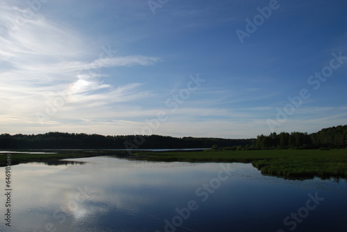 Sunset over the forest and lake. Evening landscape on the shore. The sun hangs low above the horizon, covered by high cirrus clouds. Below there is a dark forest and the calm surface of the lake.