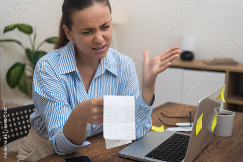 A surprised lady, homebound, confronting financial distress, holds a laptop, utility bill, and bank statements.  photo