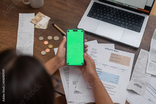 E-Paymen: A woman's hand, securing a mobile phone with a chroma key screen, rules the wooden desk surrounded by utility bills, money, and an online banking app. 