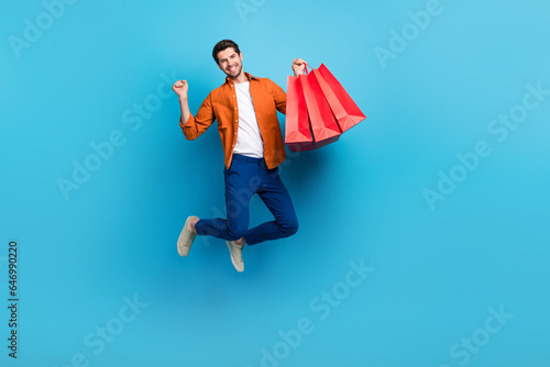 Full length photo of excited delighted man jumping hand hold boutique mall bags raise fist isolated on blue color background