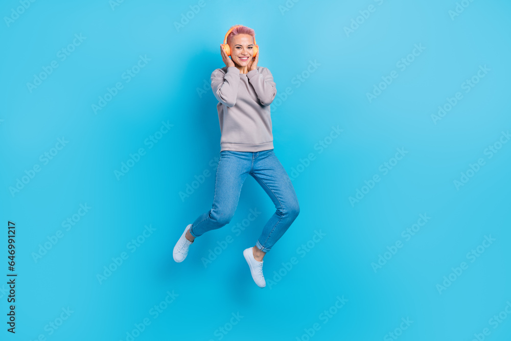 Full size photo of pretty carefree girl jumping hands touch headphones empty space isolated on blue color background Full size photo of pretty carefree girl jumping hands touch headphones empty space isolated on blue color background