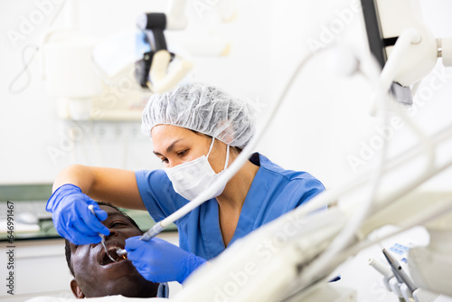 Asian woman dentist using tools for tooth repairing on african-american man patient in clinic.