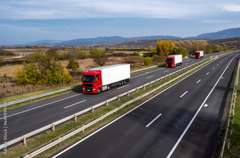 Three red trucks with a large white trailer are moving along the highway. Highway scene with Red semi trailer lorry truck passing on a highway driving at beautiful dramatic clouds. Three red trucks with a large white trailer are moving along the highway. Highway scene with Red semi trailer lorry truck passing on a highway driving at beautiful dramatic clouds.