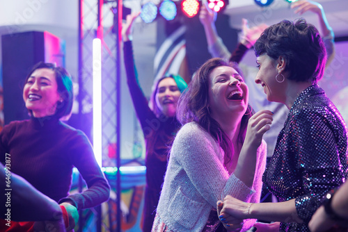 Cheerful girlfriends laughing while partying on dark dancefloor in nightclub. Happy smiling women having fun and enjoying nightlife while dancing and attending discotheque in club
