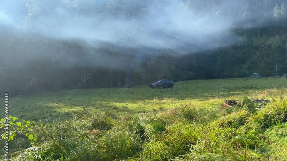 A view of a tourist campsite with tents and an electric car, where tourists are resting on a glade among trees and fog creeping over the forest. A tent city on a hike on an early sunny foggy morning. A view of a tourist campsite with tents and an electric car, where tourists are resting on a glade among trees and fog creeping over the forest. A tent city on a hike on an early sunny foggy morning.