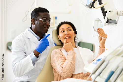 Positive asian woman and african-american man dentist looking at teeth through mirror after dental restoration procedure.