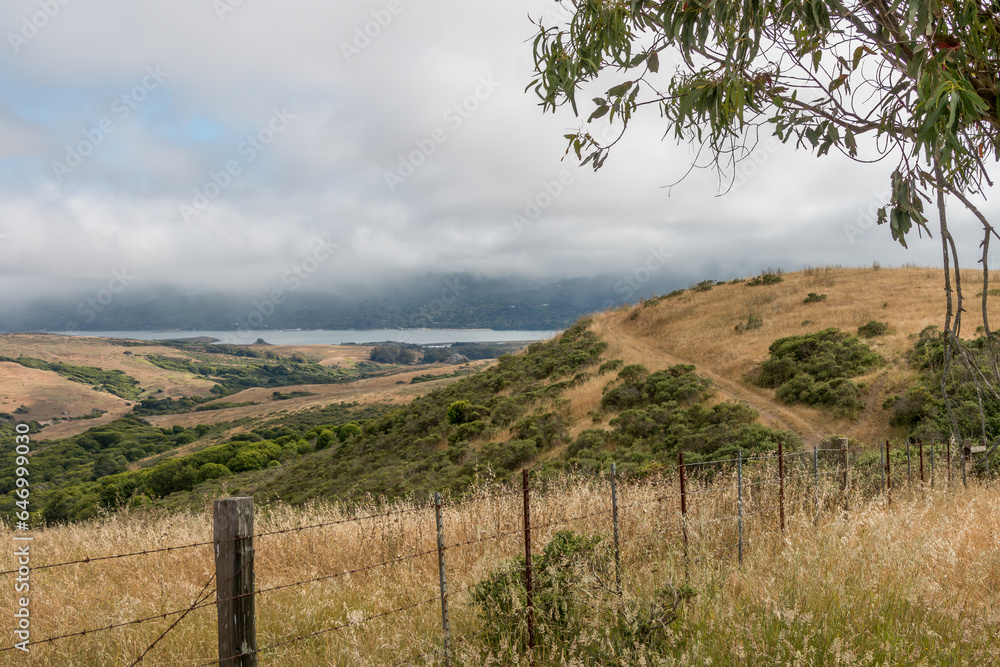 Looking over a wire fence into a golden grass valley with a lake in the distance. There is fog coming with some blue sky. Trees are in the background.