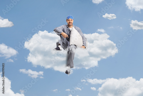 Man in pajamas with a sleeping mask holding a pillow and sitting on a cloud