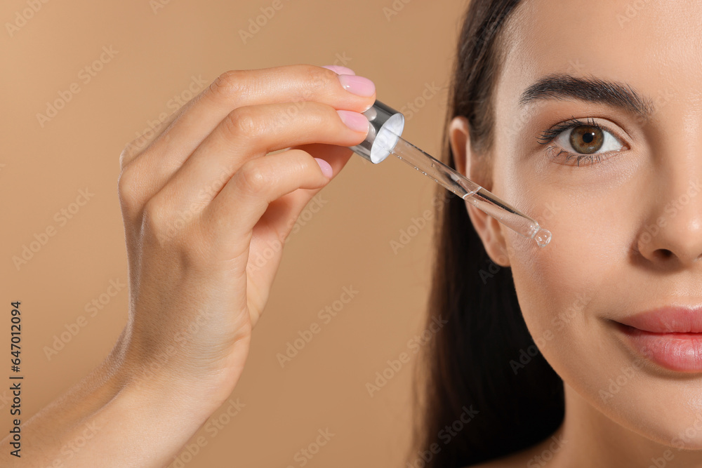Fototapeta premium Beautiful young woman applying serum onto her face on beige background, closeup