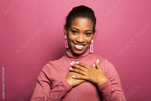 A young afro american woman with cute smile, hands near heart. Portrait on a pink background.