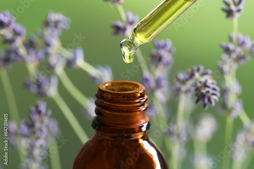 Dripping essential oil from pipette into bottle near lavender on green background, closeup