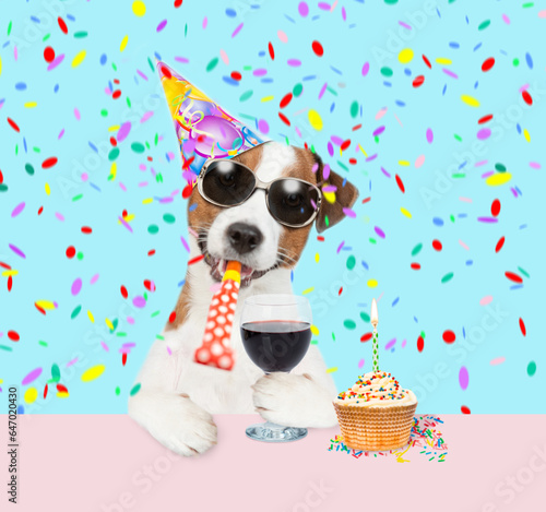 jack russell terrier puppy wearing sunglasses and party cap blows into party horn holds glass of red wine. isolated on white background © Ermolaev Alexandr