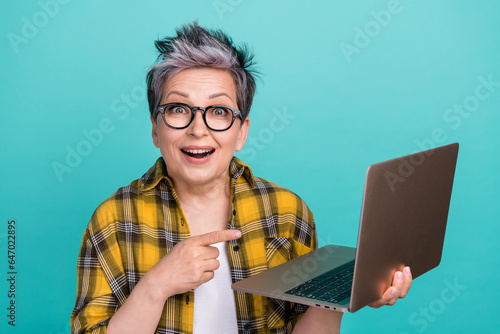 Photo portrait of lovely senior lady point empty space netbook excited wear trendy plaid yellow garment isolated on cyan color background