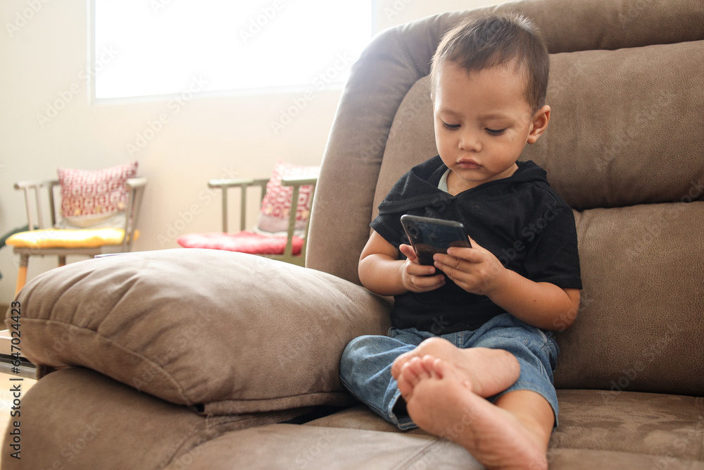 Adorable Asian boy sitting on the sofa in the living room and playing with smartphone. Adorable Asian boy sitting on the sofa in the living room and playing with smartphone.