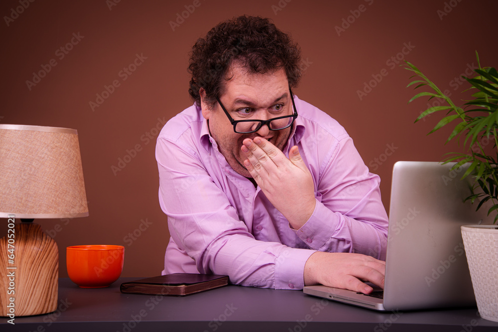 Funny fat man posing at a table with a laptop and watching adult videos. Brown background. Funny fat man posing at a table with a laptop and watching adult videos. Brown background.