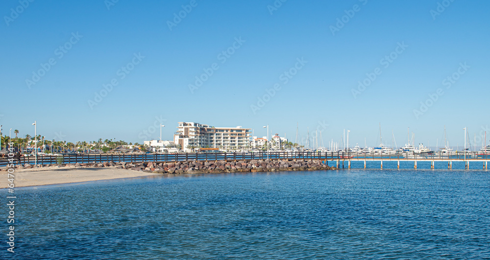 crystal clear water and a beach on the Malecon of La Paz Baja California Sur, in Mexico, with a pier in the background and a very calm sunny summer morning and clear blue sky, boats facing El Mogote. crystal clear water and a beach on the Malecon of La Paz Baja California Sur, in Mexico, with a pier in the background and a very calm sunny summer morning and clear blue sky, boats facing El Mogote.