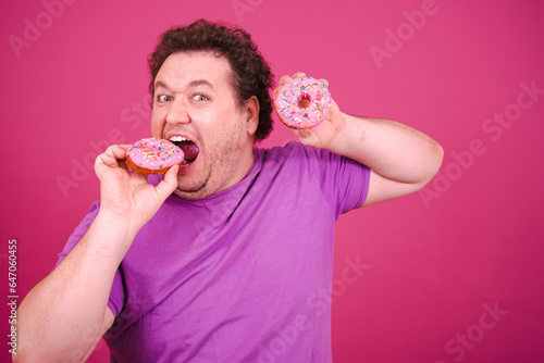 Funny fat man posing on a pink background and eating sweet donuts. Diet and healthy lifestyle.