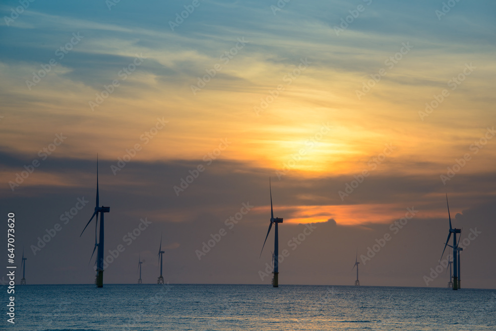 Fans of wind turbines spin over the sparkling sea. Dynamic clouds at sunset. An offshore wind farm off the northwest coast of Taiwan. One of green power.