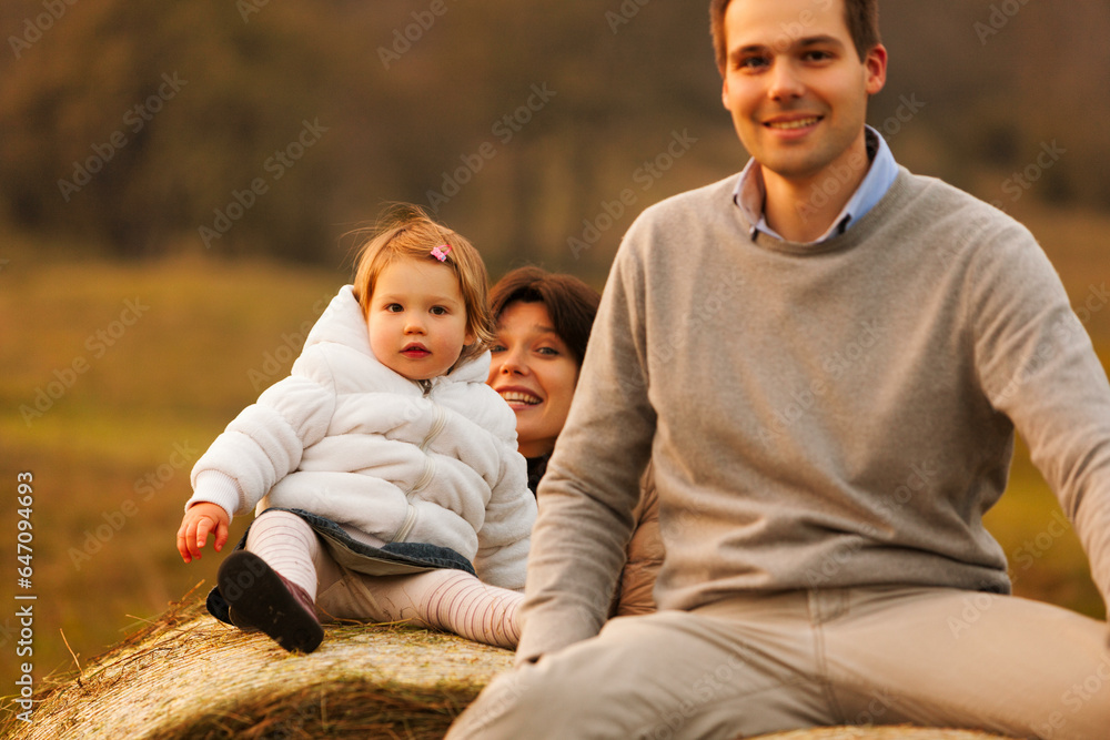 On hay bales, in the mountain foothills, loving parents let their child ascend and play. The setting sun casts a warm glow on their lively evening On hay bales, in the mountain foothills, loving parents let their child ascend and play. The setting sun casts a warm glow on their lively evening