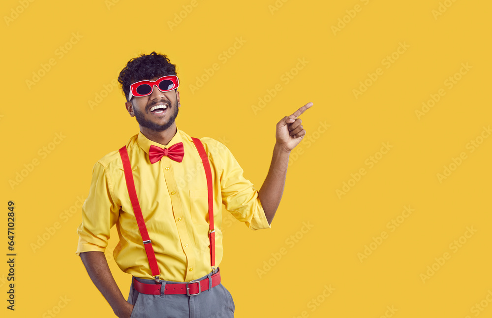 Young cheerful Indian man student in funny sunglasses for party pointing with finger at empty copy space for your promotional offer wearing flamboyant shirt stands on yellow studio background Young cheerful Indian man student in funny sunglasses for party pointing with finger at empty copy space for your promotional offer wearing flamboyant shirt stands on yellow studio background