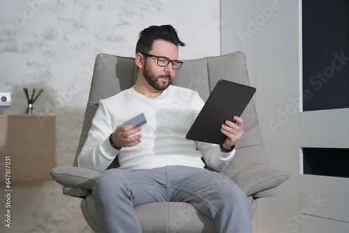 Young happy man doing online shopping with tablet laptop or smartphone, hold in hand credit bank card sitting in armchair at home in glasses 