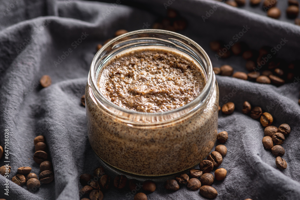 Natural coffee scrub in a transparent glass jar on the background of coffee beans on a black cloth. Concept of organic cosmetics and skin care Natural coffee scrub in a transparent glass jar on the background of coffee beans on a black cloth. Concept of organic cosmetics and skin care