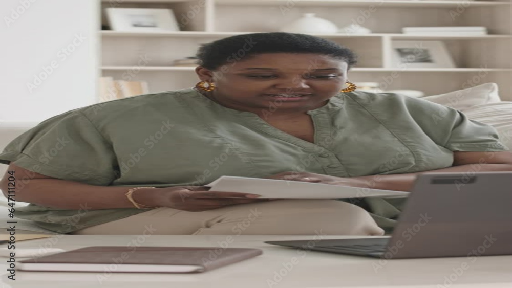 Vertical shot of black woman sitting of sofa in living room and discussing documents via online video call on laptop during e-meeting Vertical shot of black woman sitting of sofa in living room and discussing documents via online video call on laptop during e-meeting