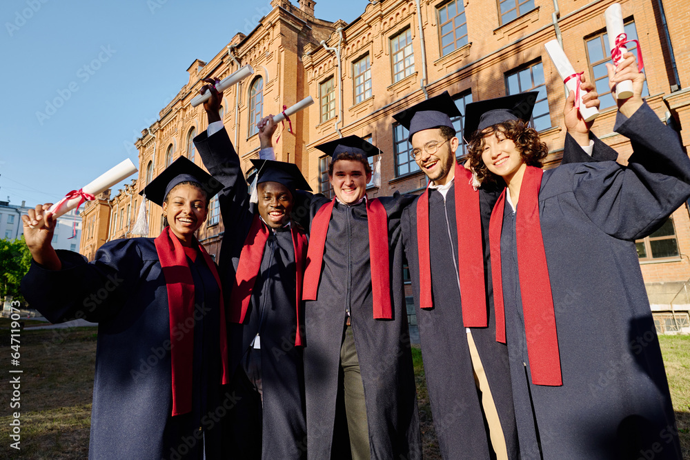 Group of happy students in gowns standing with diplomas outdoors near the university