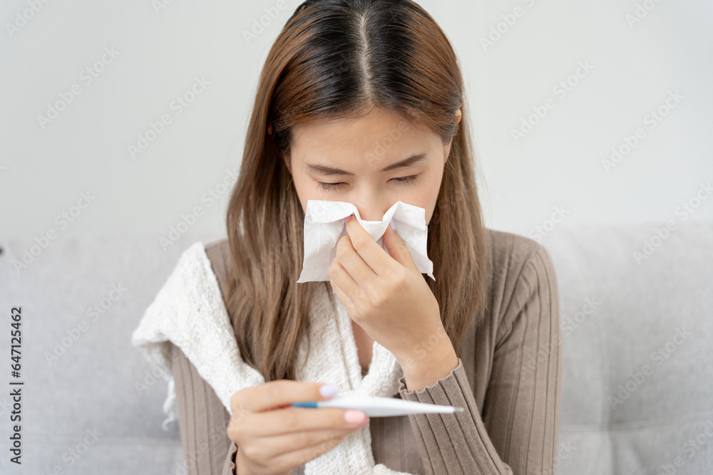 Young Asian woman having high fever while checking body temperature, female sneezing and runny nose with seasonal influenza, allergic, digital thermometer, virus, coronavirus, illness, respiratory Young Asian woman having high fever while checking body temperature, female sneezing and runny nose with seasonal influenza, allergic, digital thermometer, virus, coronavirus, illness, respiratory