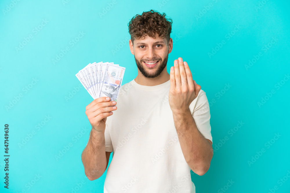 Young caucasian man taking a lot of money isolated on blue background inviting to come with hand. Happy that you came Young caucasian man taking a lot of money isolated on blue background inviting to come with hand. Happy that you came