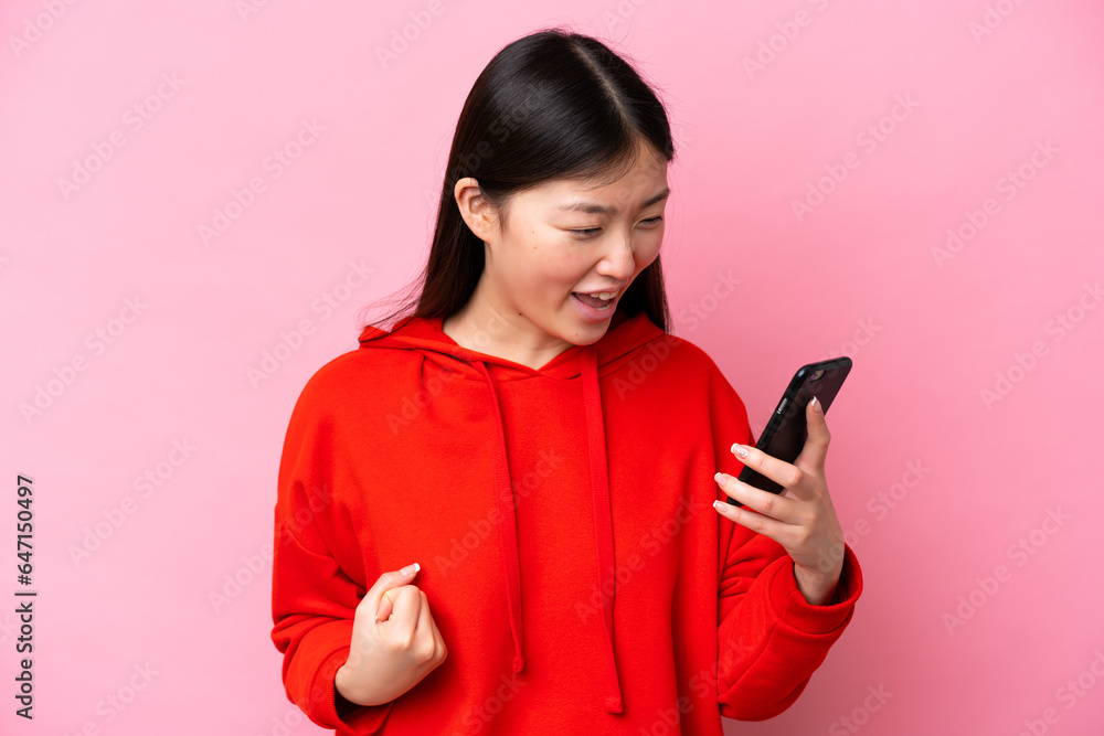 Young Chinese woman isolated on pink background using mobile phone and doing victory gesture Young Chinese woman isolated on pink background using mobile phone and doing victory gesture