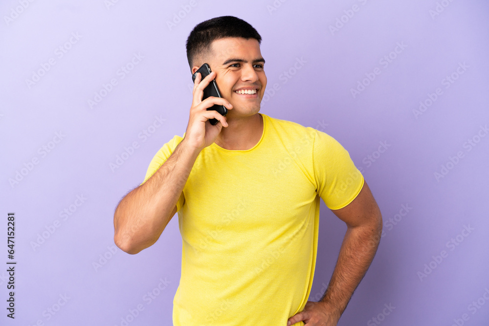 Young handsome man using mobile phone over isolated purple background posing with arms at hip and smiling Young handsome man using mobile phone over isolated purple background posing with arms at hip and smiling