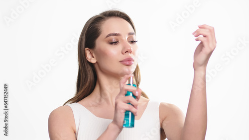 Charming beautiful young woman with light makeup holding a spray bottle and using it, isolated on white background. Concept for beauty, skin care, cosmetology, spa