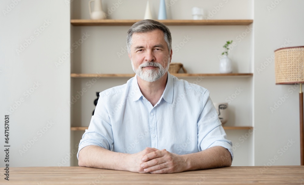 Portrait of a nice smiling grey hair man with beard, working at home, sitting at table looking at camera.