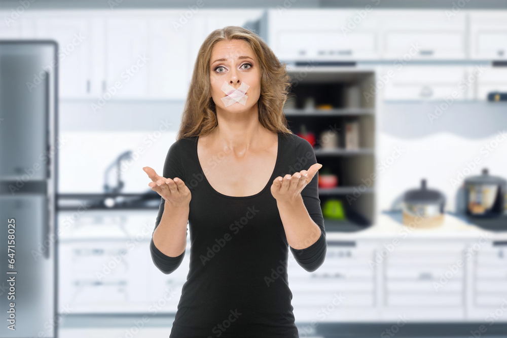 Young unhappy woman with adhesive tape on mouth posing in kitchen interior, Portrait of a displeased woman with adhesive tape in the kitchen Young unhappy woman with adhesive tape on mouth posing in kitchen interior, Portrait of a displeased woman with adhesive tape in the kitchen