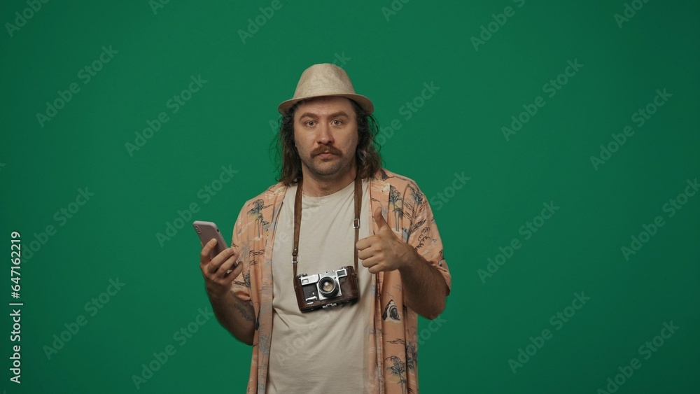Man in casual clothing and straw hat with old film camera holding smartphone, shows thumbs up, amazed face expression. Isolated on green background. Man in casual clothing and straw hat with old film camera holding smartphone, shows thumbs up, amazed face expression. Isolated on green background.
