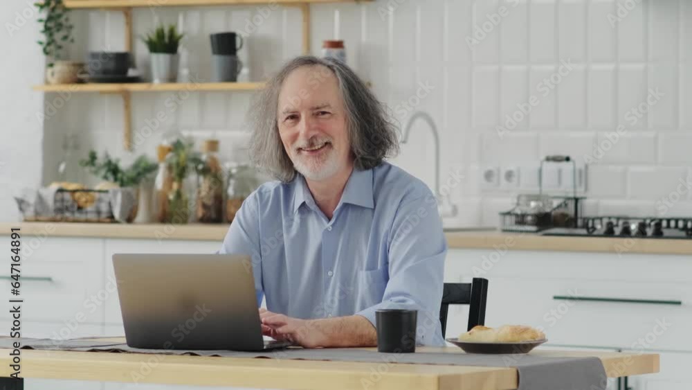 Aged remote worker. Concentrated senior male work on laptop from home office looking at the camera. Old age man employee freelancer sit at kitchen table Aged remote worker. Concentrated senior male work on laptop from home office looking at the camera. Old age man employee freelancer sit at kitchen table