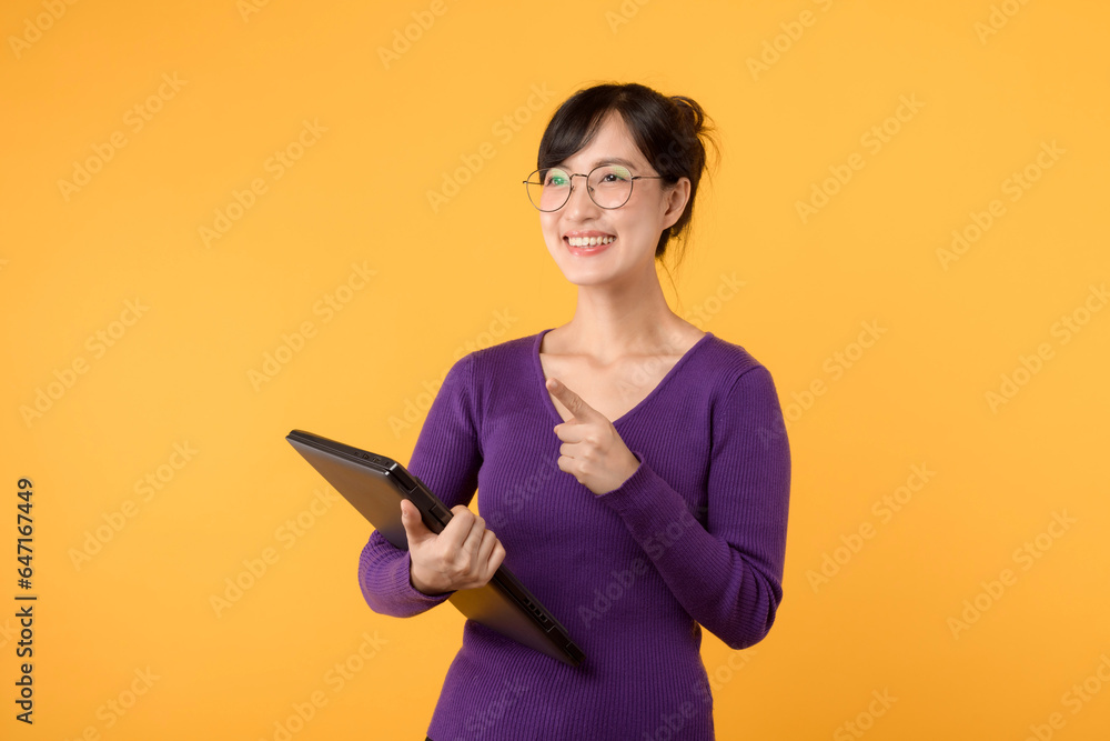 Happy asian female college student or teacher happy smiling on yellow background, holding laptop. Youth girl student pointing up to copy space. Education exchange student concept Happy asian female college student or teacher happy smiling on yellow background, holding laptop. Youth girl student pointing up to copy space. Education exchange student concept