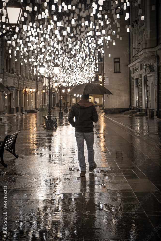 A man with an umbrella walks along a street decorated with illumination. Back view.