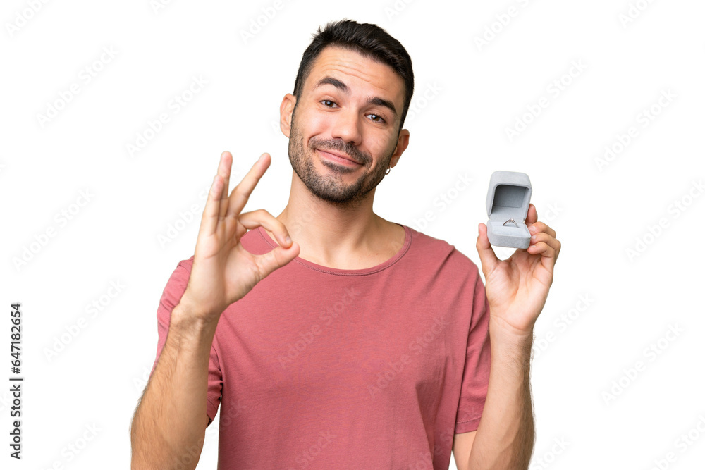 Young handsome caucasian man holding a engagement ring over isolated background showing ok sign with fingers Young handsome caucasian man holding a engagement ring over isolated background showing ok sign with fingers