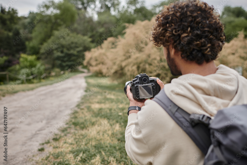 back view of curly indian photographer with backpack taking photo on professional camera outdoors back view of curly indian photographer with backpack taking photo on professional camera outdoors