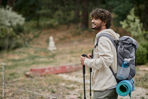 Fotografija joyful indian backpacker holding trekking poles  and looking away in forest, adv