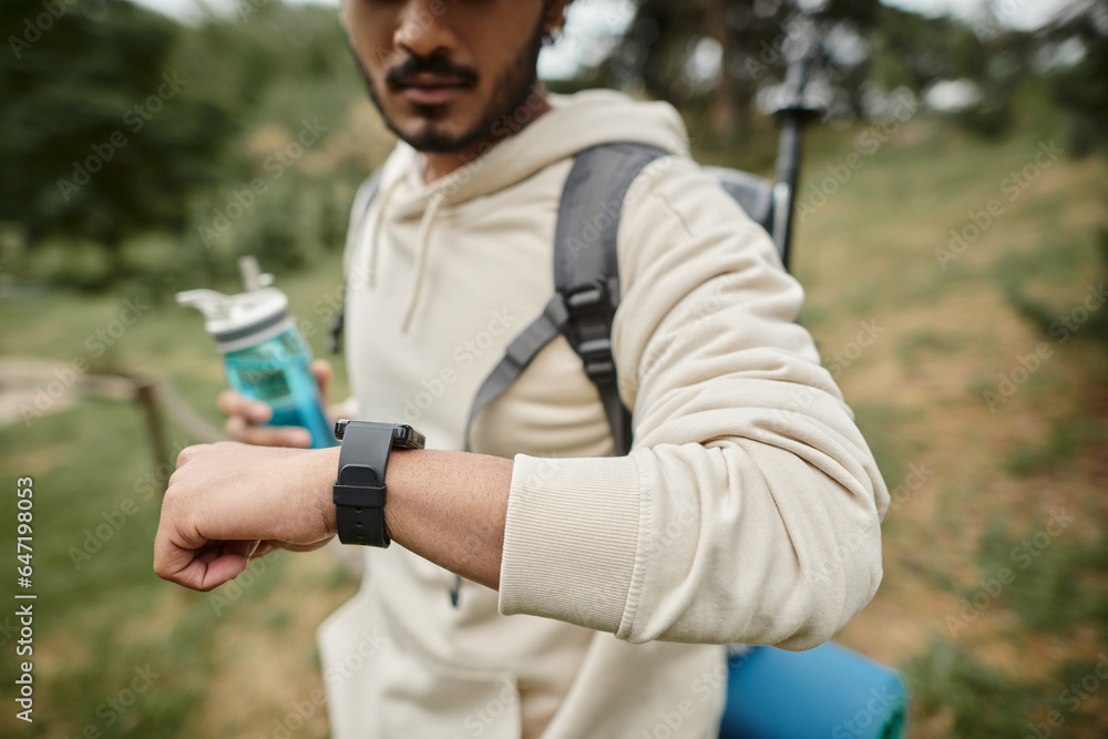 cropped view of indian tourist with backpack holding sports bottle and using smartwatch outdoors cropped view of indian tourist with backpack holding sports bottle and using smartwatch outdoors
