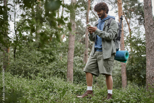 smiling young indian tourist with backpack using smartphone while standing in forest © LIGHTFIELD STUDIOS