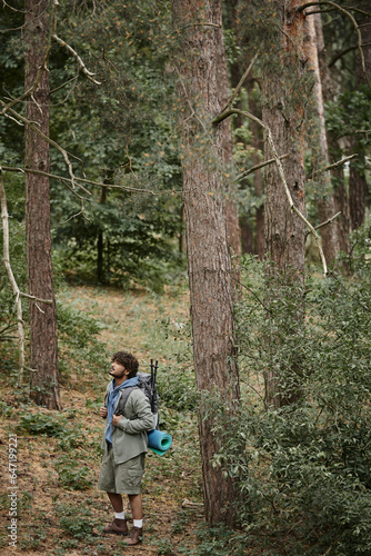 young indian man with backpack, trekking poles and fitness mat standing between trees in forest