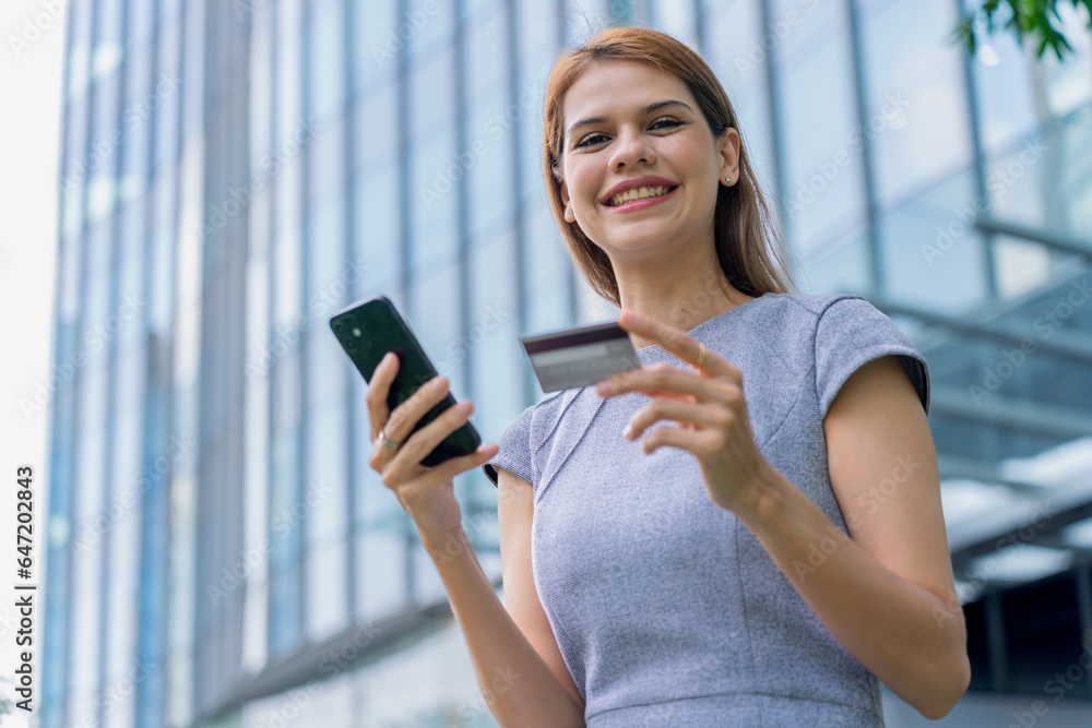 Young businesswoman doing digital banking transaction and making travel bookings using her credit card and her mobile phone in front a modern office building at a central business district